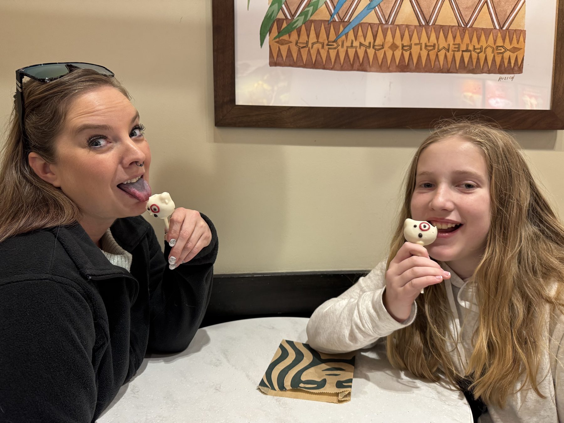 Aleasha sticking out her tongue at a cake pop across a cafe table from a laughing girl doing the same.