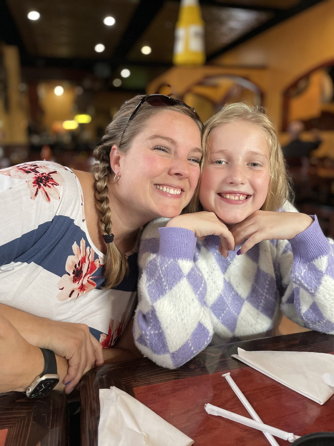 Aleasha with a braid over her shoulder leaning her head against a smiling girl in a purple sweater at a restaurant.