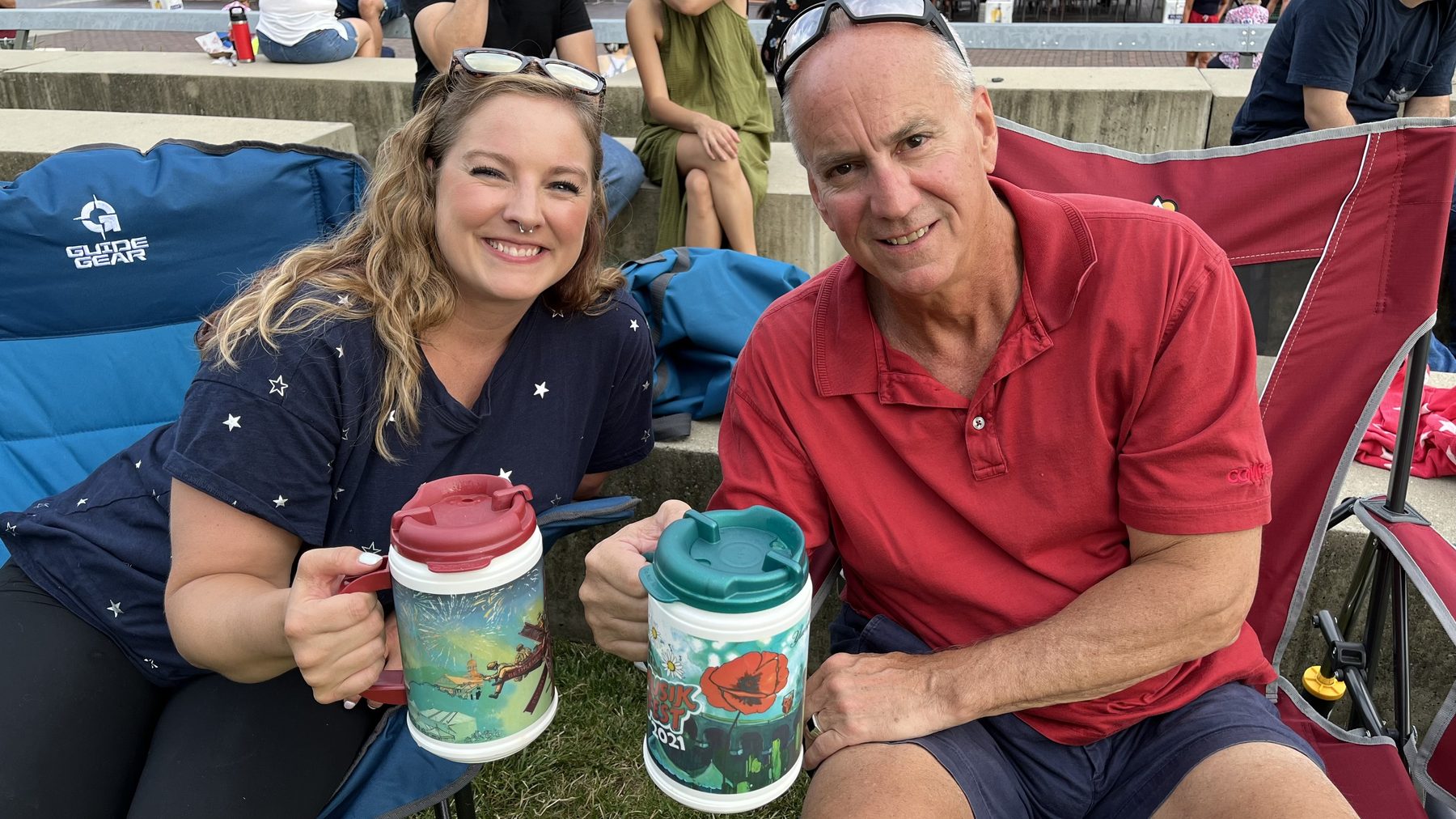 Aleasha and an older man in a red polo sitting in lawn chairs, clinking festival mugs together.