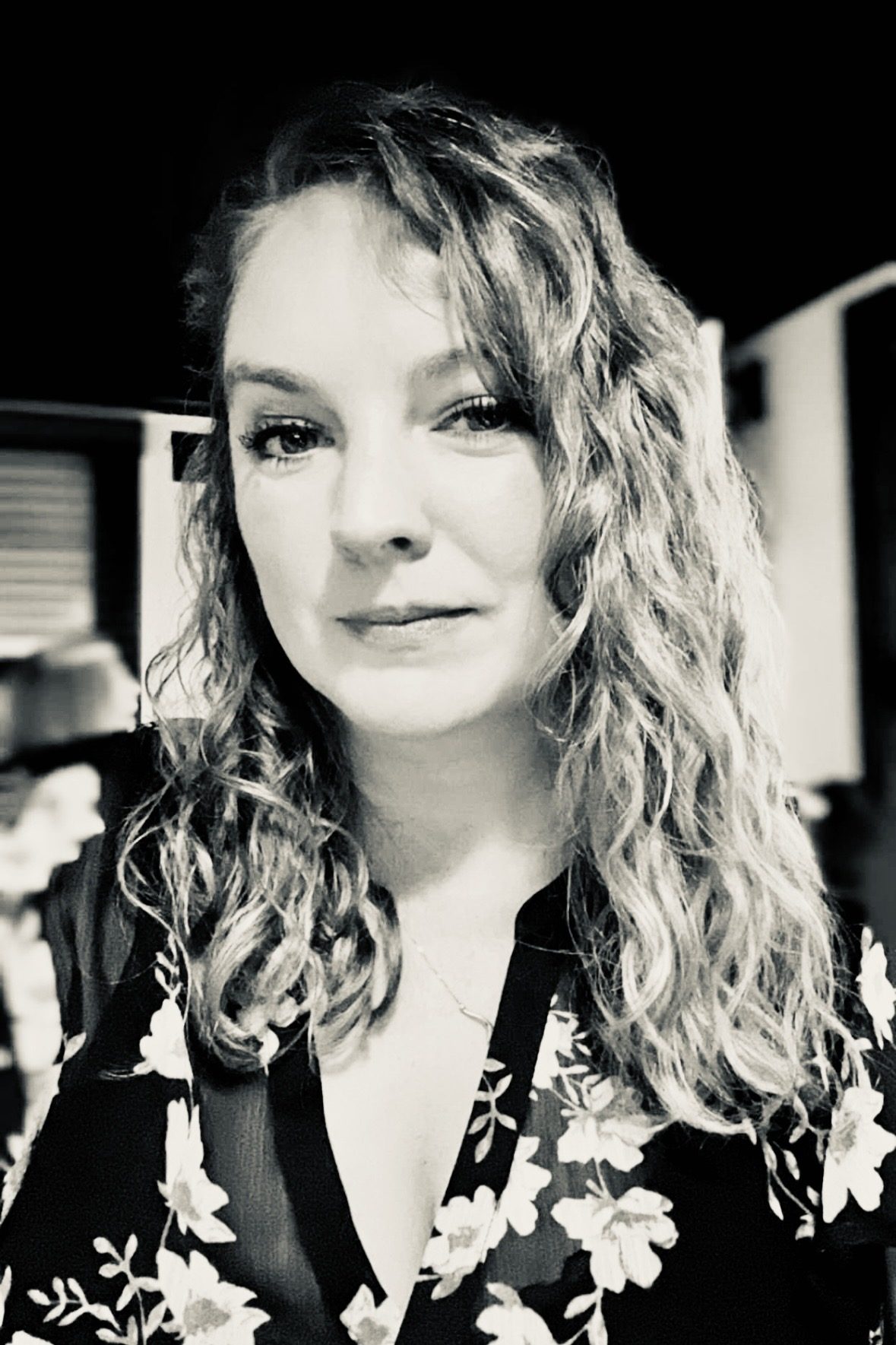 Black-and-white portrait of Aleasha with long curly hair, looking directly at the camera.